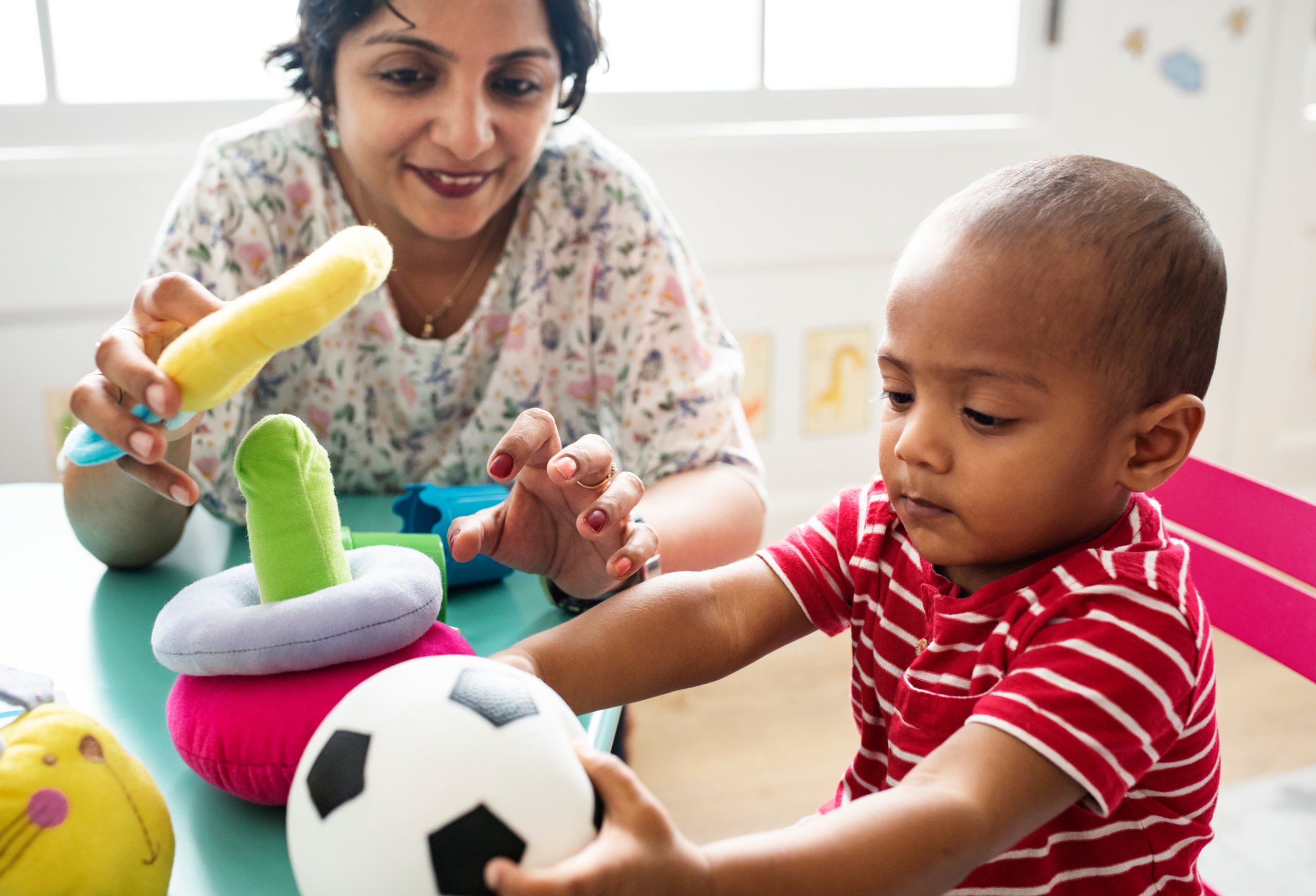 Toddler boy plays with female teacher and toys at a table.