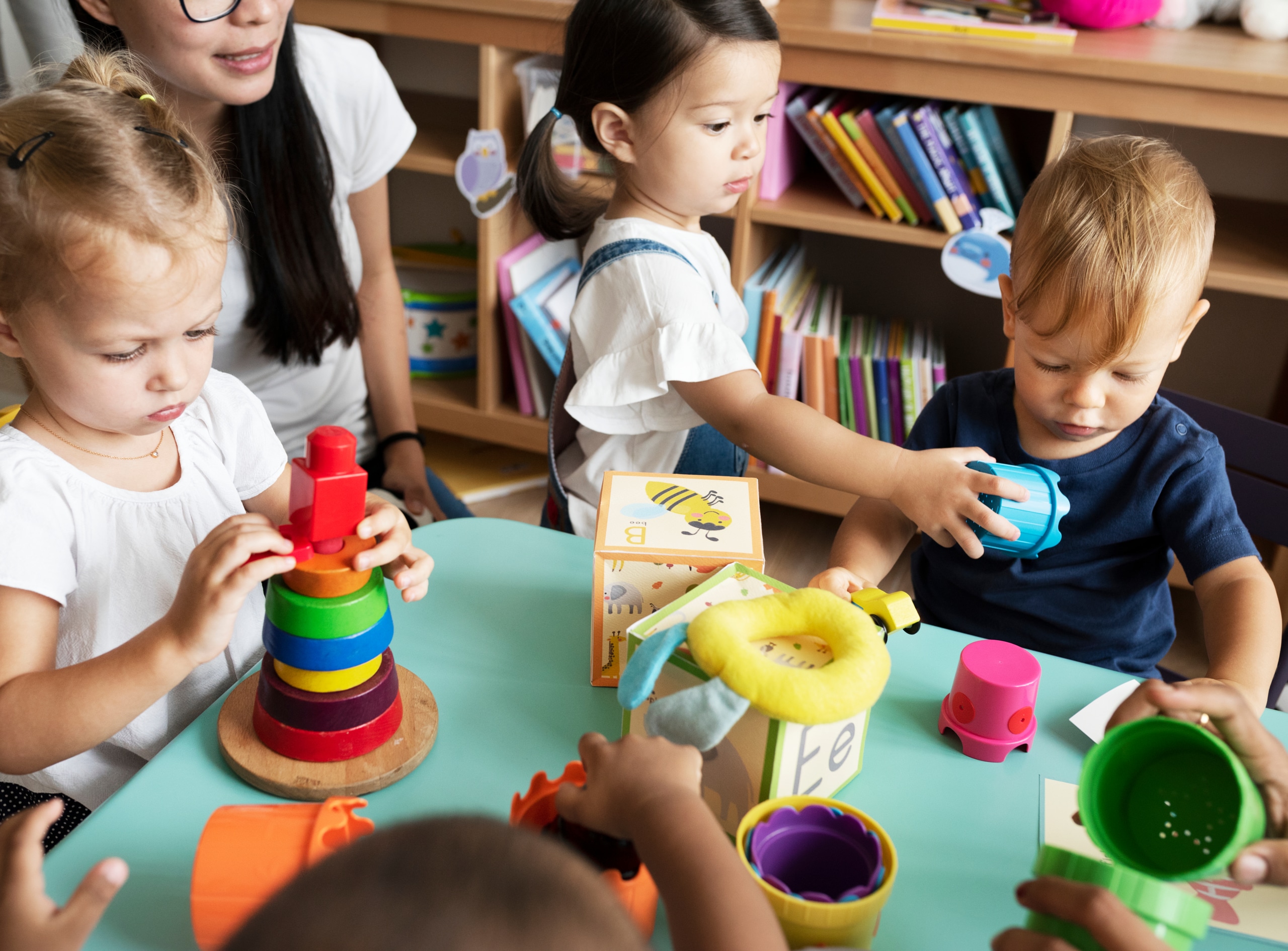 Three toddlers play with stacking toys and blocks in classroom as teacher looks on.