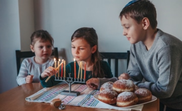 Brother and sisters siblings lighting candles on menorah for Jewish Hanukkah holiday at home. Children celebrating Chanukah festival of lights. Dreidel and Sufganiyot donuts in plate on a table.