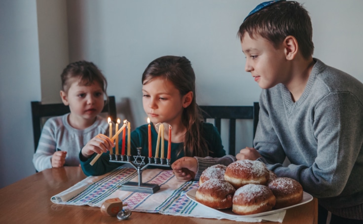 Brother and sisters siblings lighting candles on menorah for Jewish Hanukkah holiday at home. Children celebrating Chanukah festival of lights. Dreidel and Sufganiyot donuts in plate on a table.